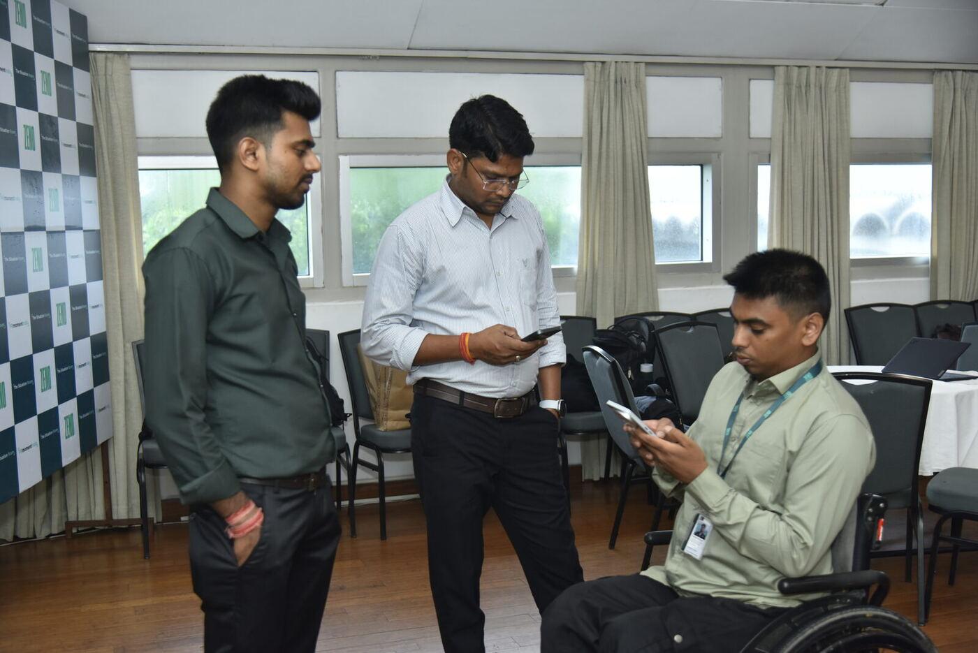 Three young delegates on their phones by the ZENO backdrop, one seated in a wheelchair
