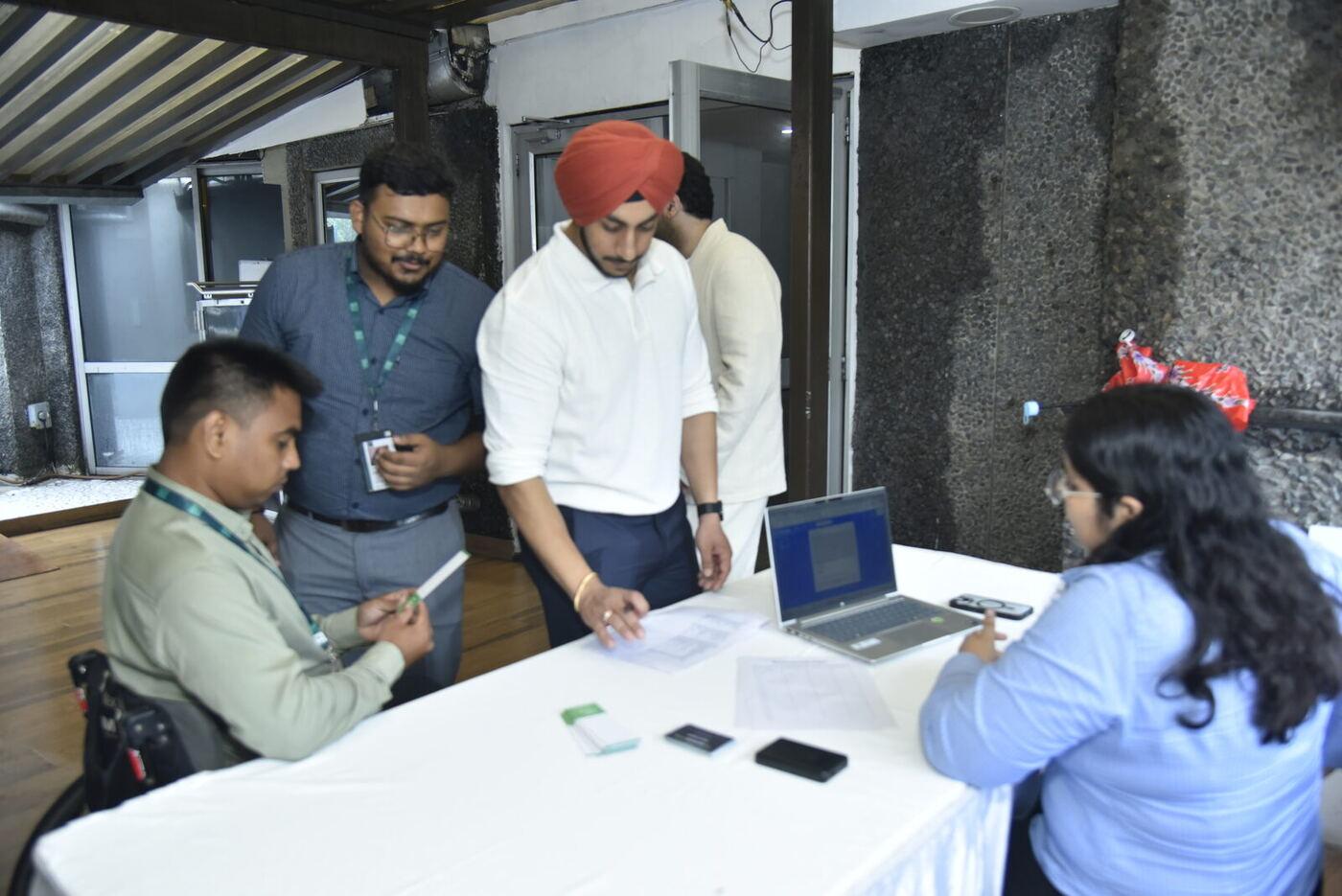 Turbaned delegate in a white polo signing the registration sheet, staff at the laptop