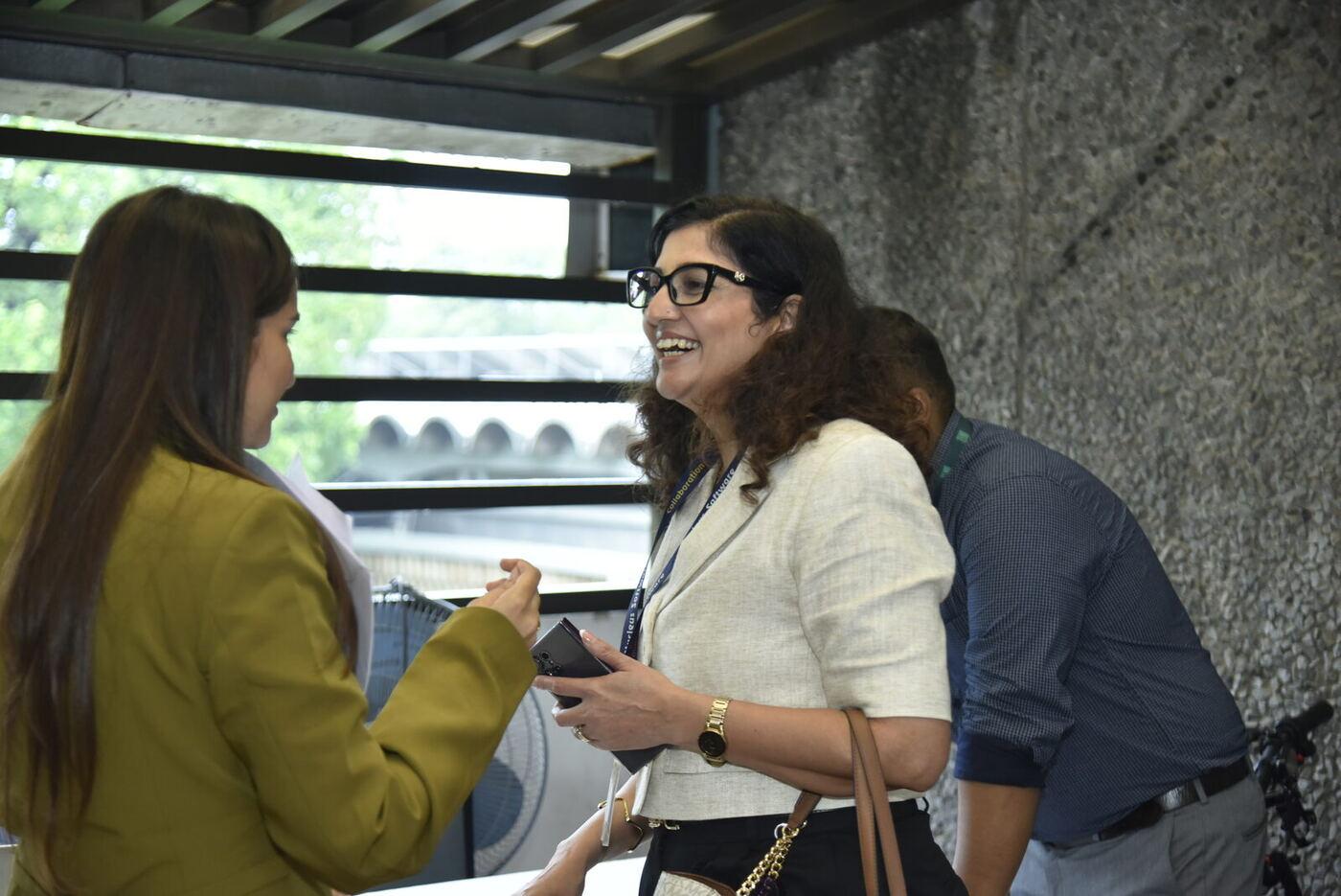 Curly-haired woman in glasses laughing mid-conversation, colleague's back to camera