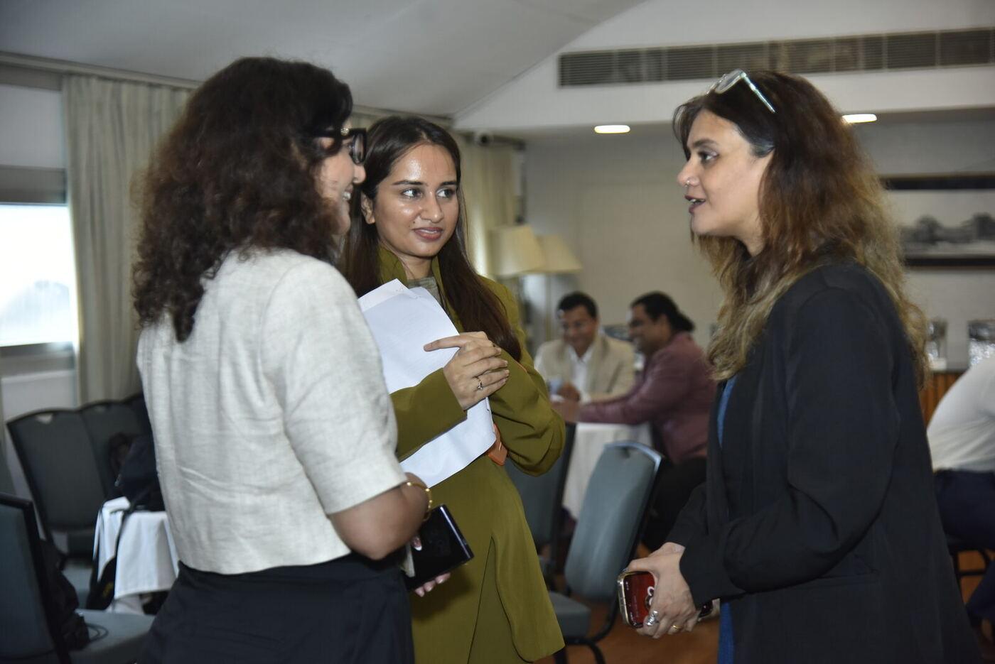 Three women standing in conversation, the one in olive gesturing with her papers