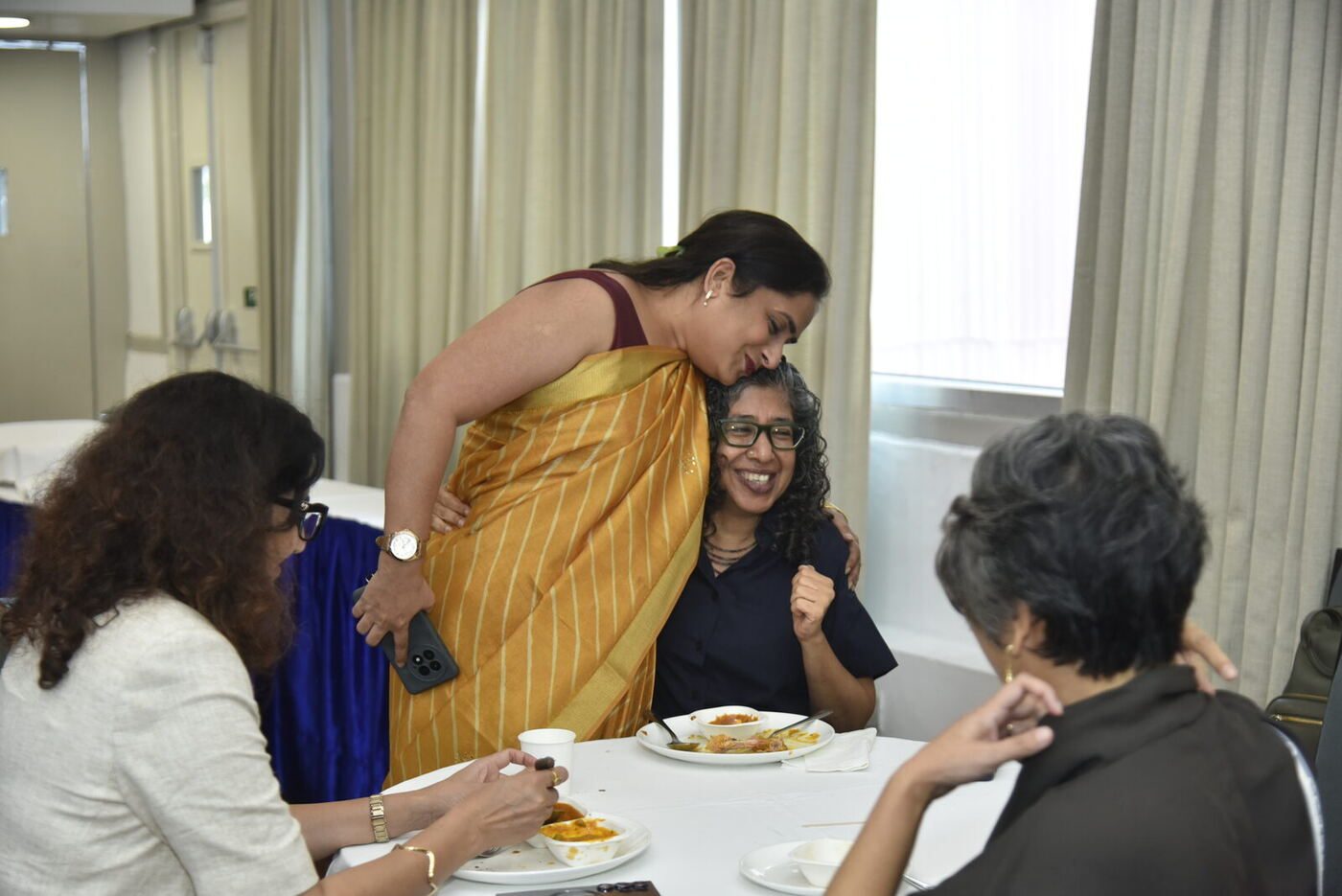 Woman in a gold saree bending to hug a seated friend, both laughing
