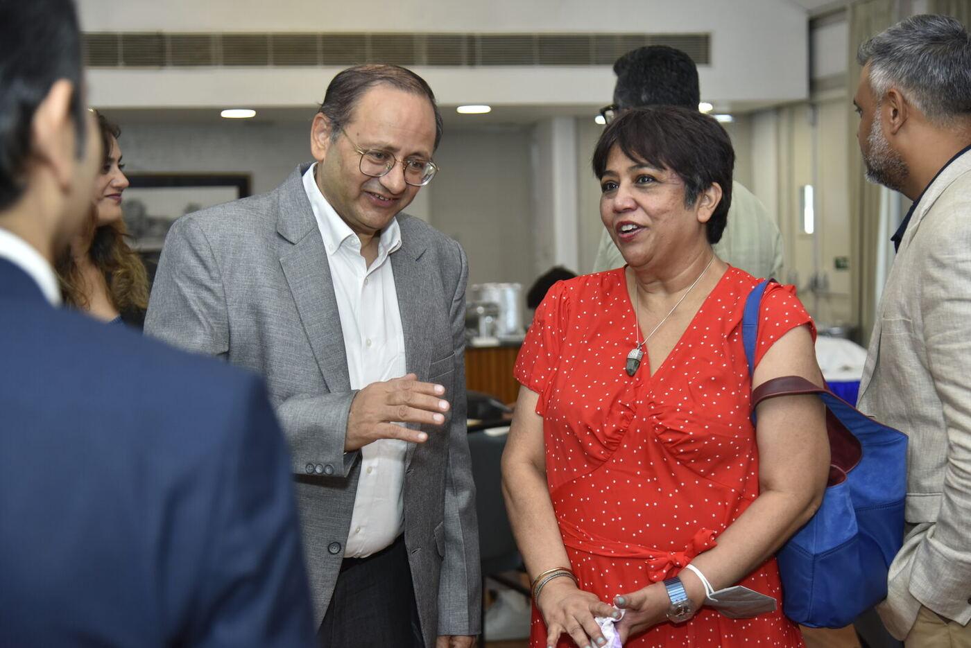 Older man in a grey blazer gesturing to the woman in red polka-dot, both smiling