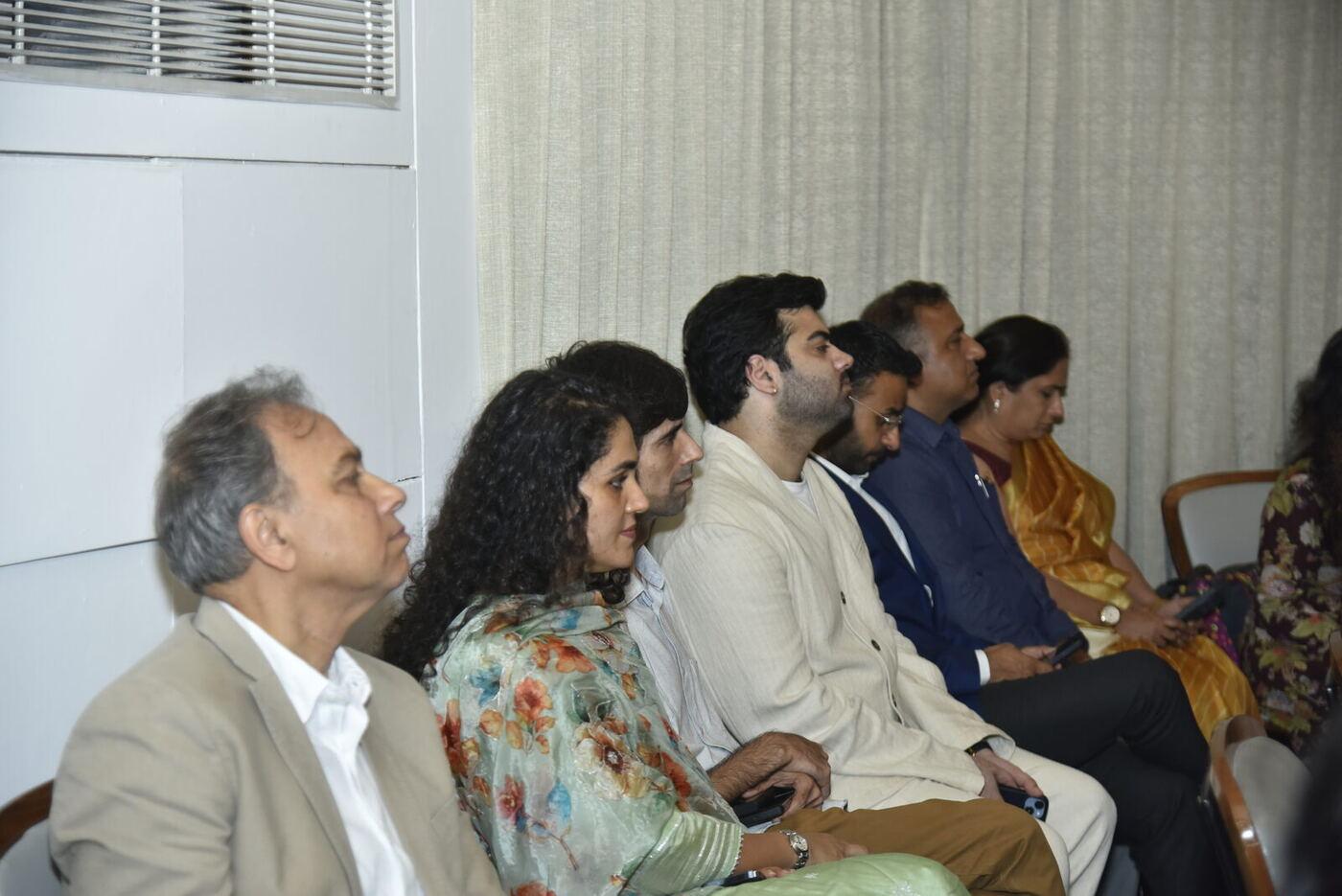 Attendee row against the curtain: older man in beige, woman in floral, man in cream kurta