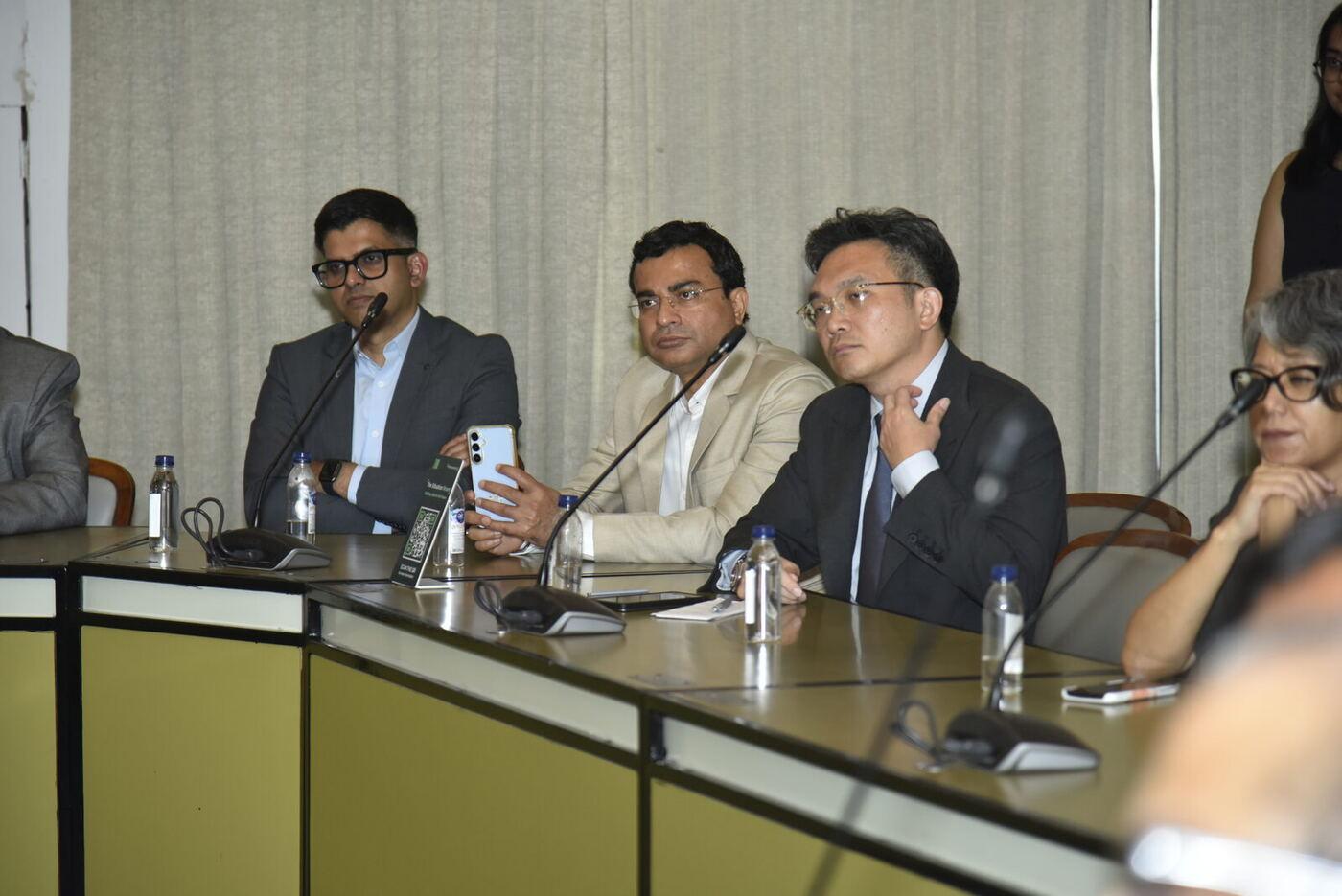 Three suited delegates, one holding his phone up to the panel, another adjusting his collar
