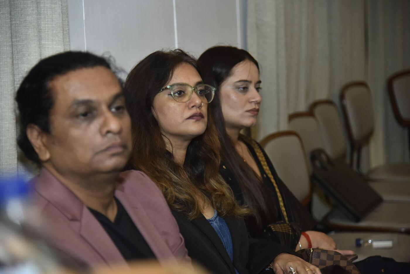Three attendees listening: man in a mauve blazer, woman in pale glasses, woman in black