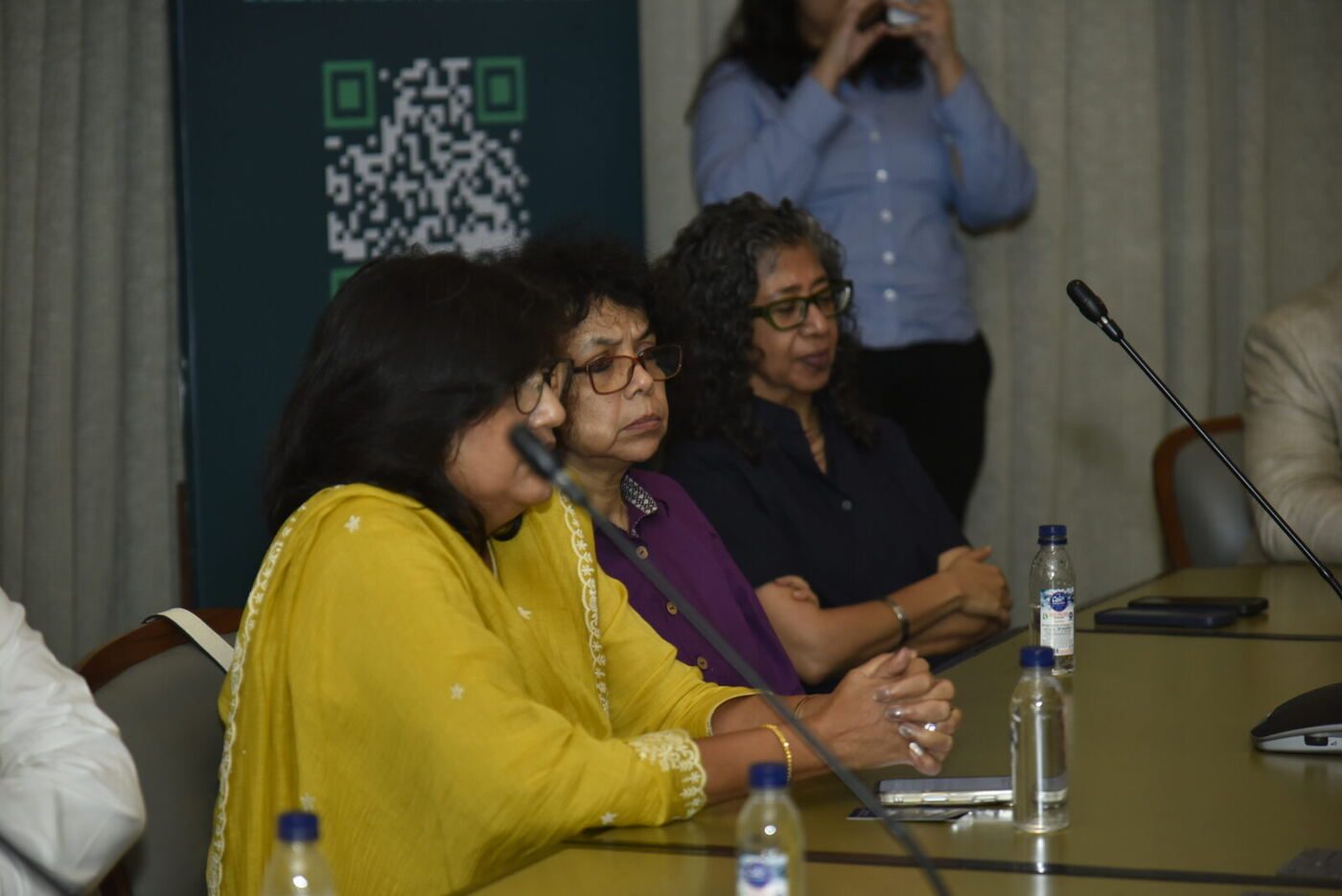 Woman in a yellow kurta, older woman in purple and woman in black glasses, all listening