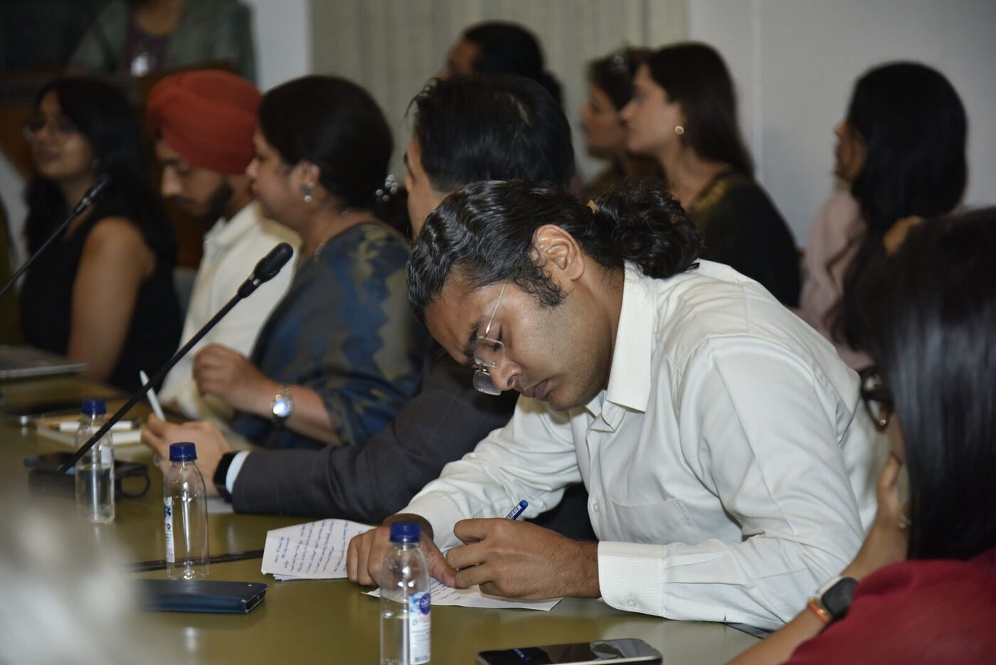 Long-haired delegate in a white shirt writing notes, others blurred behind him