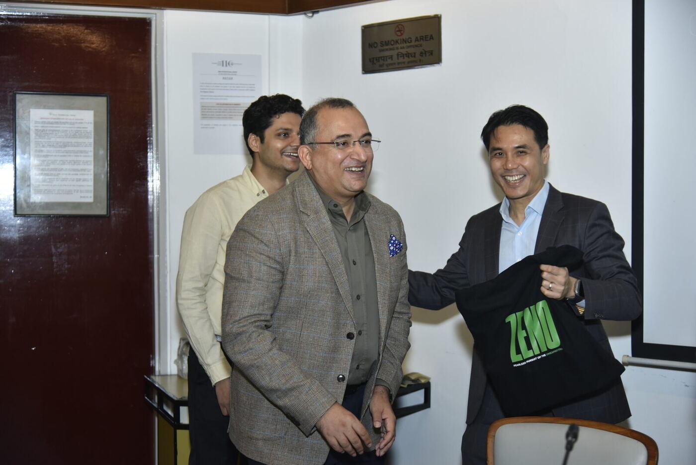 Panellist in tweed laughing as the host offers him the ZENO tote, staffer grinning behind