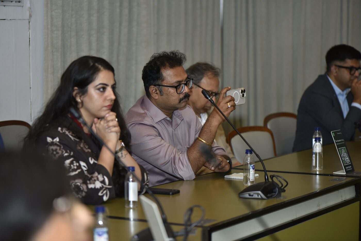 Delegate in a mauve shirt photographing the panel on his iPhone, colleague listening
